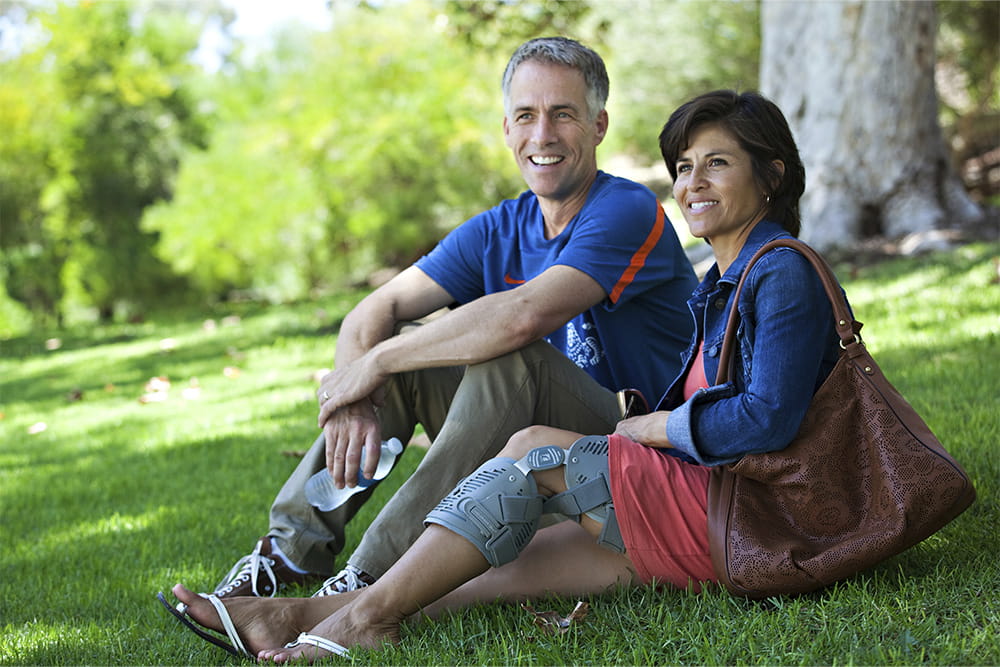 A man and a woman sit under a tree in the meadow and she wears a leg orthosis