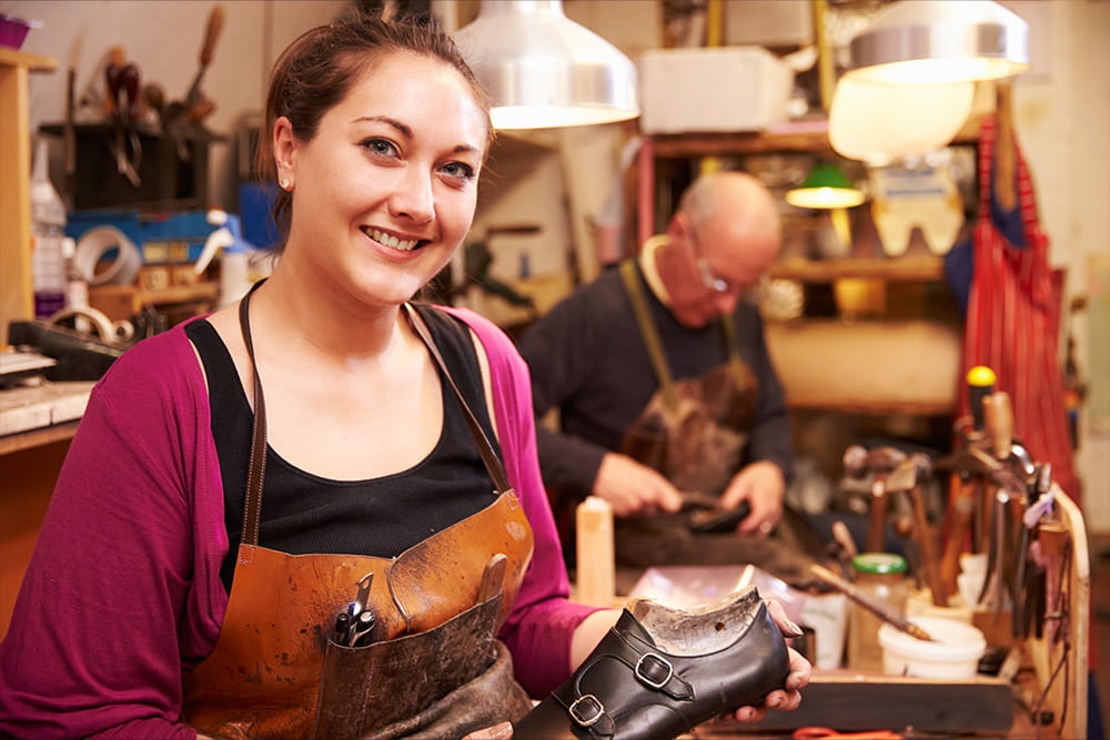 A woman in a craftsman's apron smiles at the camera and holds a shoe in her hands