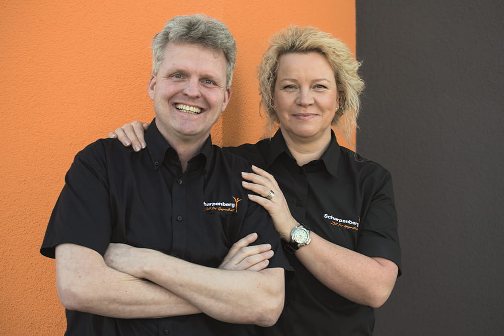 Ralph and Kathrin Scharpenberg stand in front of a house wall and laugh at the camera