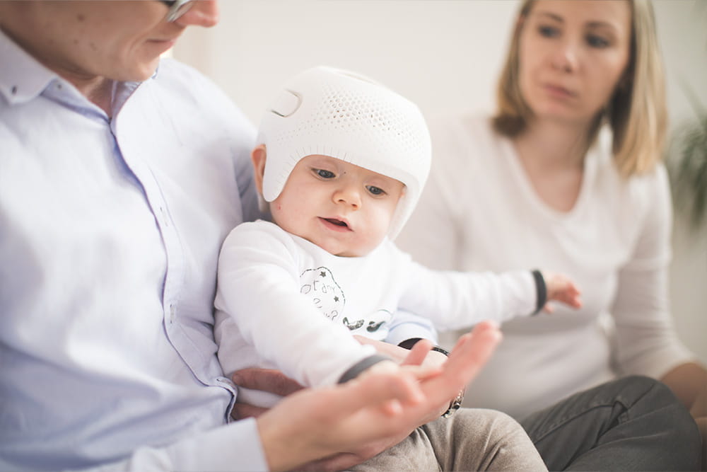 Baby mit Kopforthese sitzt im Arm des Vaters