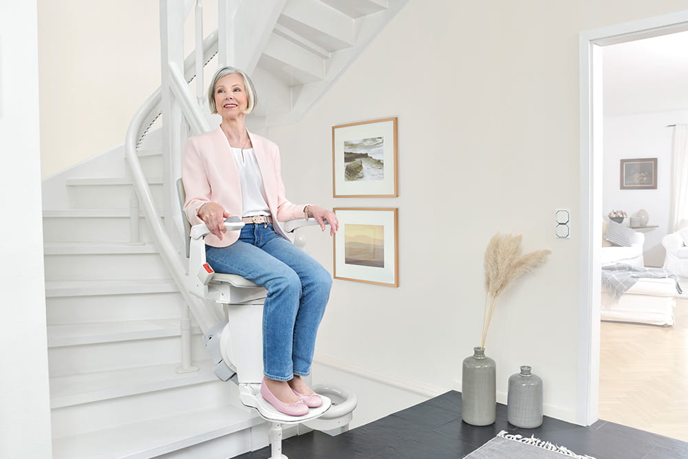 Eine Frau sitzt in einem Weiß eingerichtetem Haus auf einem Treppenlift