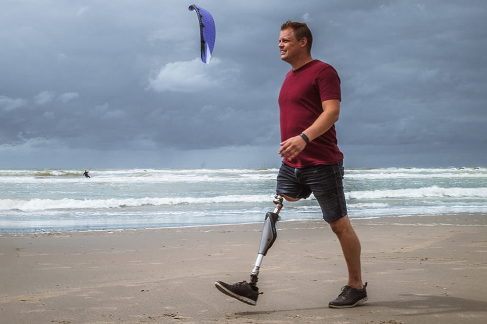 A man with endo-exo-prosthesis is walking along the beach and behind him is a kitesurfer in the water.
