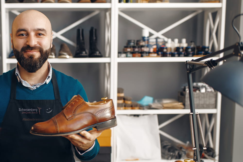 A man with a beard holds a leather shoe in his hand and smiles friendly into the camera. Shoe accessories can be seen in the background.