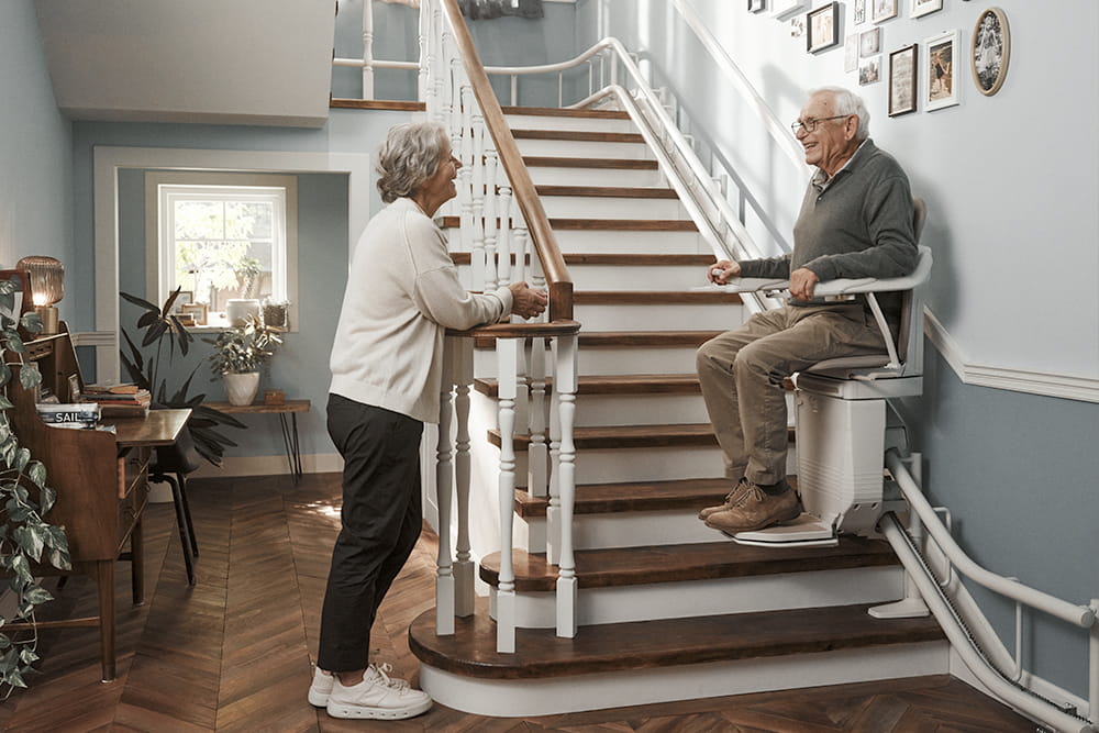Ein älterer Mann fährt gerade auf seinem Treppenlift die Treppen herunter und schaut dabei zu seiner Frau, die an der Treppe steht.