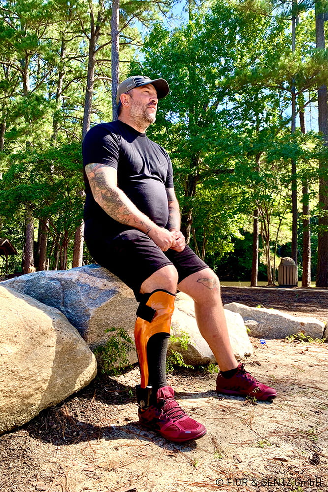 Ein Mann mit Beinorthese sitzt im Wald auf einem Stein.