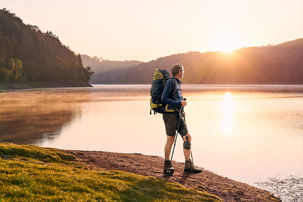 Ein Mann mit Beinorthese steht beim Sonnenuntergang an einem See in seiner Wandermontur.