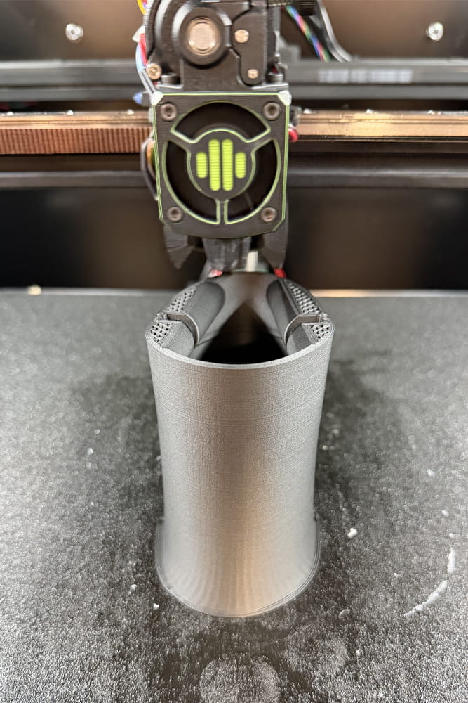 Ein 3D Drucker bei der arbeit, er druckt ein graues zylinderartiges Objekt.