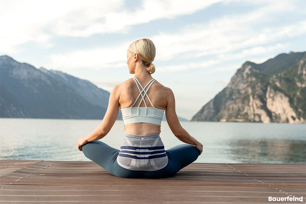 Eine Frau mit Rückenbandage sitzt auf einem Steg und dehnt sich.
